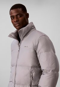 Homme portant une doudoune gris clair avec un col haut, debout devant un fond neutre uni.