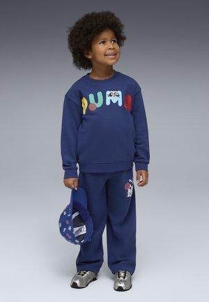 Kind trägt einen blauen PUMA-Sweatshirt und Hose mit bunten Buchstaben und Basketball-Motiven, hält eine passende blaue Mütze, blickt nach oben und lächelt.