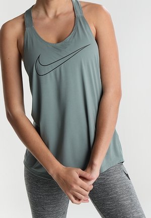 Femme portant un débardeur Nike gris ample et des leggings de sport gris, debout, les mains jointes devant un fond uni.