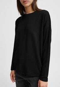 Maglietta nera a maniche lunghe con una texture a costine, scollatura rotonda e vestibilità rilassata. Abbinata a jeans grigio scuro, che mette in evidenza la semplicità del capo.