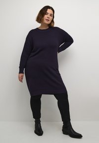 Dark purple, ribbed knit dress with long sleeves, a round neckline, and dropped shoulders, paired with black fitted pants and ankle boots.