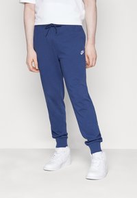 Modré bavlněné tepláky s elastickými manžetami a pasem, s malým bílým logem. Kombinovány s bílými vysokými teniskami.