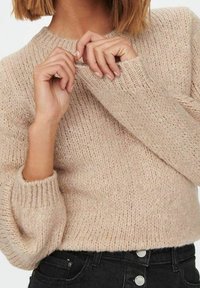 Donna che indossa un maglione beige a maglia con polsini e colletto a costine, che aggiusta il neckline, abbinato a jeans neri a vita alta.