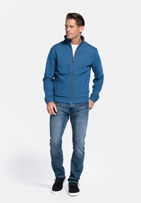 Blauwe zip-up jack met staande kraag, textiel met structuur, gecombineerd met lichtblauwe jeans en zwarte sneakers.