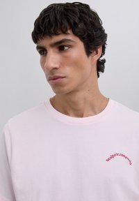 Młody mężczyzna z ciemnymi, kręconymi włosami, ubrany w jasnoróżową koszulkę z czerwonym napisem "scalpers company" na piersi, spogląda w bok.