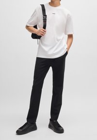 Biała bawełniana koszulka z logo "HUGO", połączona z czarnymi spodniami i czarnymi sneakersami. Model trzyma czarną torbę na ramię.