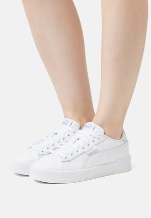 Weiße Puma Low-Top-Sneaker, getragen an nackten Beinen vor einem schlichten hellen Hintergrund, die Seiten- und Vorderansichten beider Schuhe zeigen.