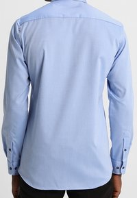 Camicia azzurro chiaro a maniche lunghe con una texture liscia, colletto appuntito, orlo curvo e bottoni neri sui polsini e sul collo.
