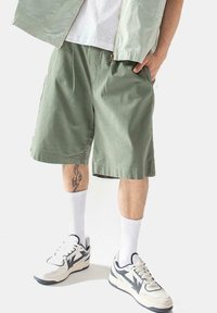 Pantalones cortos de algodón verde con un corte holgado, bolsillos laterales, usados con calcetines blancos y zapatillas gruesas. Tatuaje visible en la pierna.