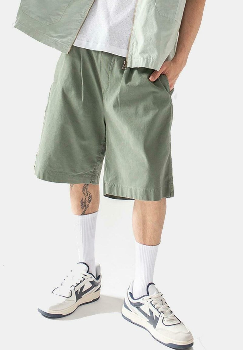 Pantalones cortos de algodón verde con un corte holgado, bolsillos laterales, usados con calcetines blancos y zapatillas gruesas. Tatuaje visible en la pierna.