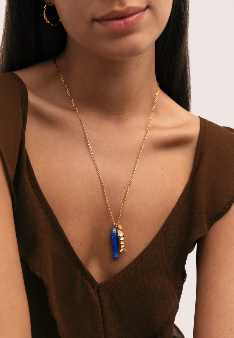 Mujer con collar de cadena dorada con colgantes de pez azul y espina de pescado dorada, combinado con pendientes de aro dorados y top marrón con cuello en V.