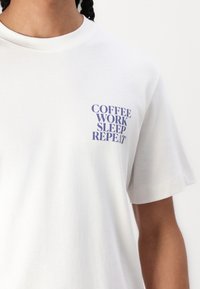 Weißes Baumwoll-T-Shirt mit kurzen Ärmeln. Mit blauer Schrift: "KAFFEE ARBEIT SCHLAF WIEDERHOLEN" auf der linken Brustseite.