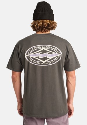 Person med krøllet hår iført en sort beanie og en mørkegrå T-shirt med stort "Billabong Boardshorts" logo på ryggen.