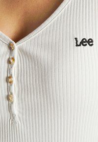 Bílý žebrovaný oděv s výstřihem do V a čtyřmi kulatými knoflíky. Na levé straně je černě vyšitý nápis "Lee". Měkká textura.