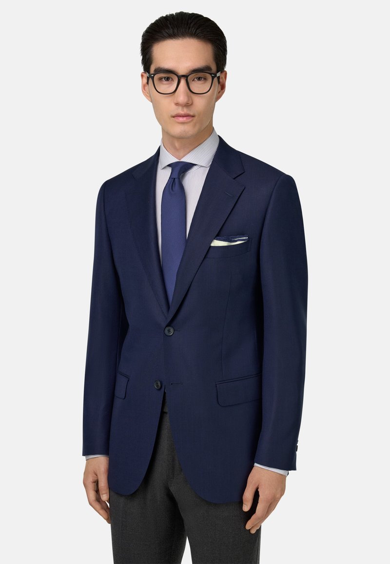 Námornícky modrý oblekový sako z textúrovanej látky, s klopovými reverzami, dvoma gombíkmi a bočnými vreckami, v kombinácii s ľahko pruhovanou košeľou a kravatou.