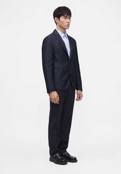 Homme debout face à l’objectif, portant un costume foncé avec une chemise bleu clair et des chaussures noires, sur fond blanc uni.