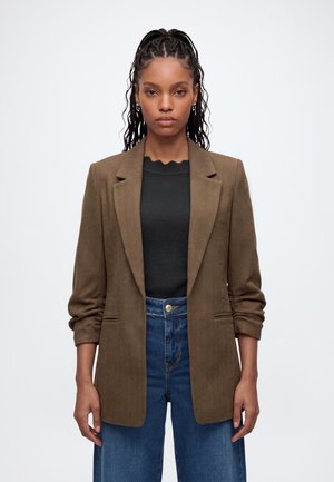 Femme aux longs cheveux tressés portant un blazer marron sur un top noir et un jean bleu taille haute, debout devant un fond blanc.