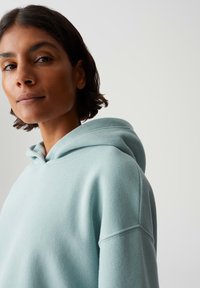 Donna con i capelli scuri e corti che indossa una felpa con cappuccio di un blu chiaro, che guarda leggermente in alto e di lato contro uno sfondo chiaro e semplice.