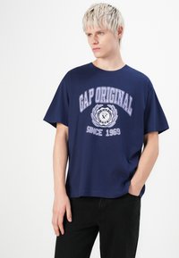 Sötétkék pamut póló rövid ujjakkal, "GAP ORIGINAL" felirattal és logóval fehér és világoslila színben, laza fit.
