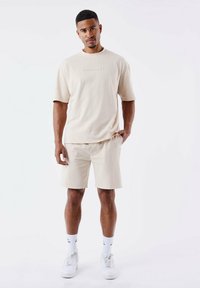 T-shirt in cotone beige a maniche corte con un piccolo logo sul petto, abbinata a pantaloni corti beige, indossata con sneakers bianche e calzini a fantasia.