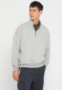 Sudadera gris con cremallera y collar alto, puños y dobladillo acanalados. Presenta un sutil logo en el pecho y un material suave y texturizado.