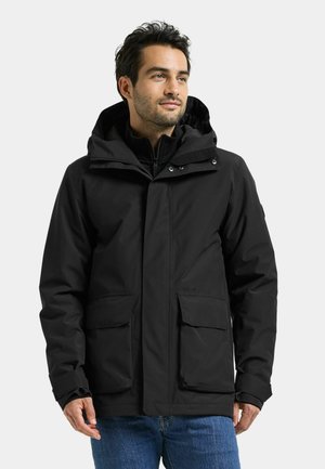 Mann trägt eine schwarze Winterjacke mit Kapuze und vorderen Klappentaschen sowie blaue Jeans, schaut leicht zur Seite.