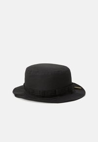 Chapeau de seau noir avec un bord étroit et des détails cousus, arborant une petite étiquette sur le côté.