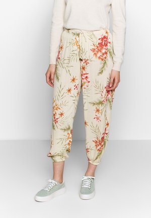 Frau trägt cremefarbene, mit Blumen bedruckte Hose mit Kordelzug und hellgrüne Sneakers, steht vor einem schlichten hellen Hintergrund.