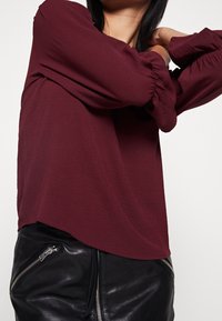 Bordeaux pitkähihaisessa paidassa on smokkikuitut, ja se on valmistettu kevyestä, teksturoidusta kankaasta. Yhdistetty mustiin nahkahousuihin, joissa on vetoketjuyksityiskohtia.