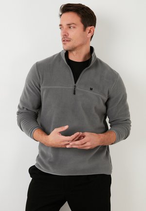 Pullover grigio in pile con colletto a zip a un quarto, dotato di un piccolo logo nero, maniche lunghe e una texture morbida.