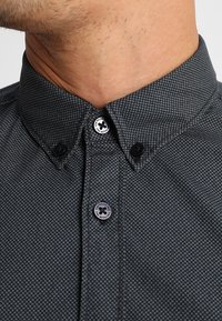 Kortärmad, donkergrijze shirt met een subtiele textuur en een klein wit stippenpatroon. Voorzien van zwarte knopen, een klassieke kraag en merkknopdetail.