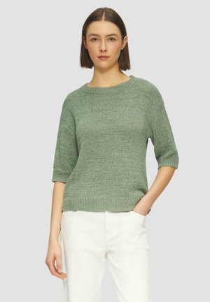 Maglioncino verde a maglia con scollatura rotonda, maniche corte e orlo a costine. Indossato con pantaloni bianchi, presentato su uno sfondo neutro.