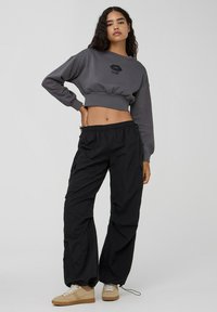 Wycięta szara bluza z czarnym odciskiem ust, połączona z luźnymi czarnymi spodniami i beżowymi tenisówkami z białymi akcentami.