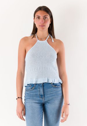 Giovane donna con un top a collo alto di maglia azzurro chiaro e jeans blu, in piedi davanti a uno sfondo bianco semplice.