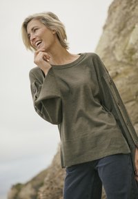 Pull en maille olive avec un col large, des manches surdimensionnées et des boutons sur le côté, associé à un jean bleu foncé. Tissu texturé.