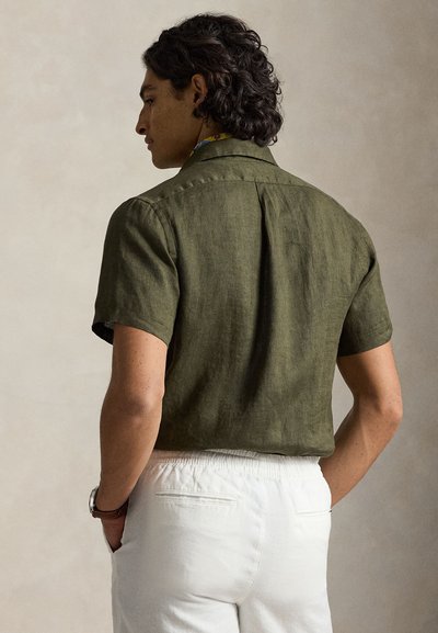 Άνδρας με ελιάς πράσινο κοντομάνικο πουκάμισο και άσπρο παντελόνι, στέκεται με τα χέρια στις τσέπες, στραμμένος προς τα αριστερά σε μονόχρωμο φόντο.