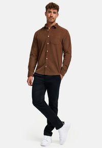 Barna, mintás anyagból készült gombos ing, egy mellzsebbel, fekete nadrággal és fehér tornacipővel párosítva.