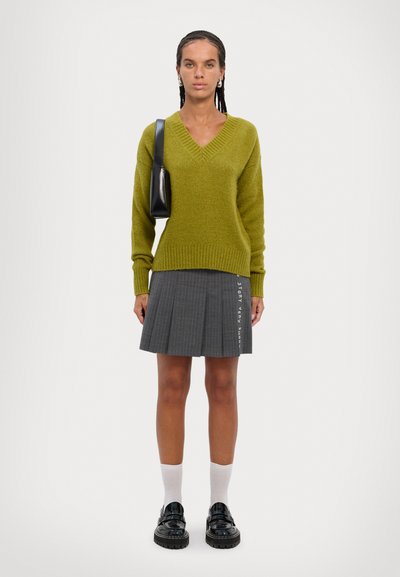Pulover verde tricotat cu decolteu în v, fustă plisată gri cu text, șosete albe și pantofi negri. Design simplu, texturi moi și culori contrastante.