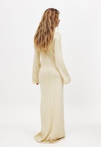 Robe longue beige en tissu doux, dotée d'une silhouette cintrée, de manches longues et de coutures visibles le long du dos.