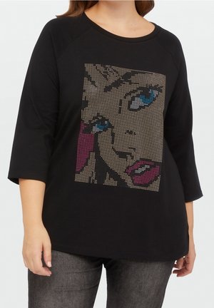 Femme portant un haut noir à manches trois-quarts avec un visage pop art pixellisé en bleu, beige et rose sur le devant.