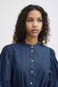 Denim bluza s visokom izrezom, napuhanim rukavima i dugmadi sprijeda. Boja je srednje plava s mekanom teksturom i suptilnim sjajem.