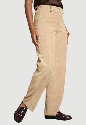 Person trägt beige Cordhosen mit weitem Bein, dunkelbraune Loafers, ein dunkles Oberteil und eine goldene Armbanduhr mit Ring an der linken Hand.