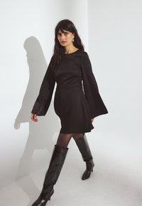 Robe noire en satin avec de longues manches évasées, taille cintrée et jupe évasée. Associée à des bottes noires à talons aiguilles montantes et à des collants transparents.