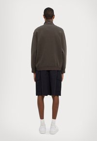 Felpa verde scuro con colletto alto, abbinata a pantaloni cargo blu navy. I pantaloni presentano tasche laterali e sono lunghi fino al ginocchio. Sneakers bianche completano l'outfit.