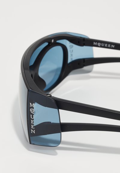 Ochelari de soare cu ramă neagră și lentile nuanțate de albastru. Prezintă un design curbat și eticheta "MQUEEN" pe latura ramei.