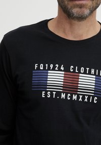 Juodas ilgomis rankovėmis marškinėliai su grafiniu spausdiniu, kuriame yra horizontalių dryžių mėlyna, raudona ir balta spalvomis, taip pat tekstas, kuris skelbia "FQ1924 CLOTHING EST. MCMXXIC."