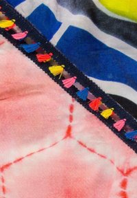 Kolorowa tkanina w niebieskie i żółte wzory; posiada czarną lamówkę z wielokolorowymi frędzlami (różowe, niebieskie, żółte, czerwone) wzdłuż krawędzi.