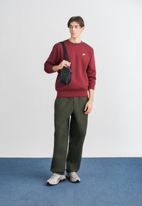 Červená mikina, zelené cargo nohavice, šedé tenisky. Čierna crossbody taška. Model stojí na svetlom pozadí na modrej podlahe.