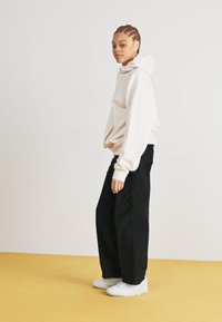 Ανοιχτό μπεζ φούτερ με καγκουρό τσέπη και υπερμεγέθη εφαρμογή, συνδυασμένο με μαύρο παντελόνι σε ίσια γραμμή και λευκά αθλητικά παπούτσια, σε κίτρινο δάπεδο.