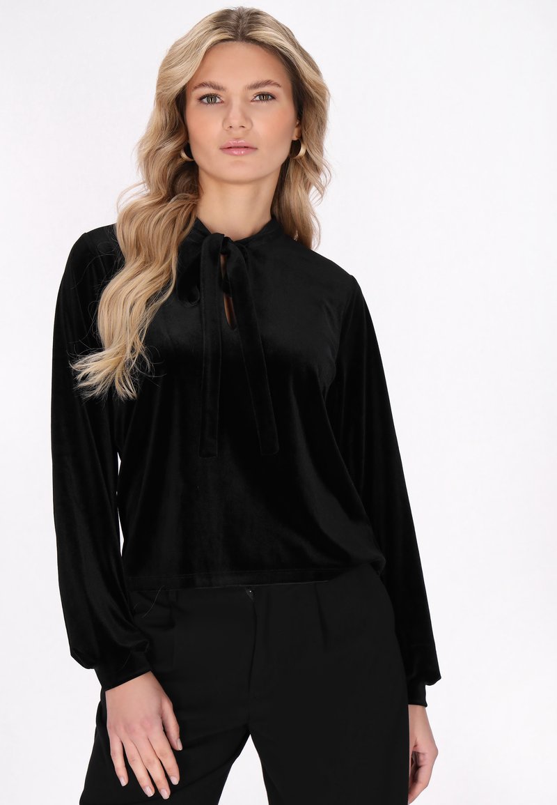 Blusa in velluto nero con maniche lunghe, colletto alto e chiusura con nodo al collo. Abbinata a pantaloni neri. Texture liscia.